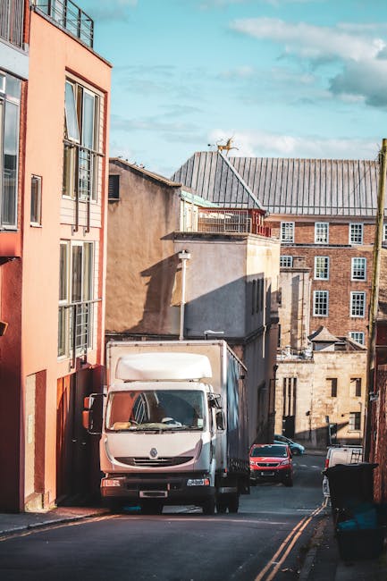 A white delivery van is parked on a narrow urban street, positioned close to the curb near a building with red and beige exterior walls. The van's front is facing the camera, and it is situated on the road with two yellow roadside markings visible. Behind the van, a small red car is parked facing away, and another vehicle is partially visible further down the street. Several old brick and concrete residential buildings with various window styles and rooftop structures are visible in the background. The street appears to be part of a home relocation or moving process, with the van ready for loading or unloading furniture or packing materials. Bright daylight illuminates the scene, with shadows cast by the buildings and the vehicles, indicating clear weather. This setting reflects typical urban moving logistics, with furniture transport prepared for quick loading and secure transport by Kingston Removals, supporting seamless house removals in Kingston.
