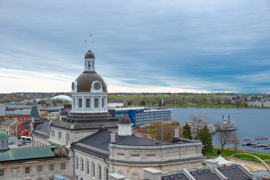 Aerial view of a historic building with a large clock tower featuring a rounded dome and small windows, situated in an urban area near a river. Surrounding the building are various structures with rooftops made of metal and tiles, some with chimneys. In the foreground, there are rooftops with flat and sloped surfaces, one with a skylight, and a green corrugated metal roof. The setting shows a cityscape with a mix of modern and older architecture, a river with boats docked along its banks, and a pathway with trees and lawns nearby. The scene is captured during daytime with overcast skies, providing natural, diffused lighting. The context hints at a city centre possibly involved in relocation or home moving activities, with parts of the scene suggesting a loading area or transportation hub, supported by the proximity to river transport and urban infrastructure, which Kingston Removals might use for furniture transport and home relocation services.