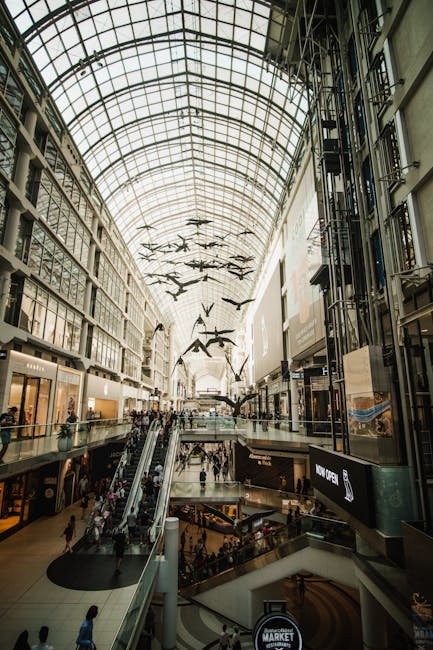 Interior view of the Bentall Centre Kingston shopping mall, featuring a high, curved glass roof supported by metal framework allowing natural light to illuminate the space. Multiple floors with retail stores are visible, connected by glass-fronted elevators and escalators. Inside, shoppers are walking through wide corridors, some on the ground and upper levels. Hanging from the ceiling are decorative, abstract bird-shaped sculptures. Packaging materials such as cardboard boxes, plastic wrap, and packing blankets are seen stacked on trolleys and near the entrance area, indicating ongoing packing and furniture transport activities. Kingston Removals staff may be involved in the loading process, although no movers are visible. The scene demonstrates a busy retail environment with elements consistent with logistics and moving services related to property and shop fitout relocation projects, as referenced on the Kingston removals website under the 'removals' service category.