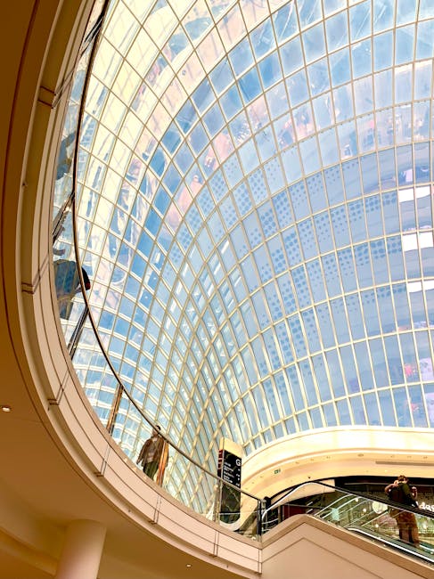 Interior view of the Bentall Centre Kingston, showing a large, curved glass skylight ceiling with a grid pattern of metal frames, allowing natural light to illuminate the shopping mall. Below the skylight, a white circular balcony with a metal handrail extends around the upper level, with a few shoppers present. On the lower level, a section of the shopping mall can be seen, along with some retail signage and decorative elements. The scene captures the architectural design of the shopping centre, emphasizing the spaciousness and modern glass construction typical of retail and shop fitouts in Kingston, with visible activity related to moving furniture or packaging materials occurring near the balcony edge. Kingston Removals would facilitate the logistics and transport of retail equipment within such a property during a shop fitout or relocation process.