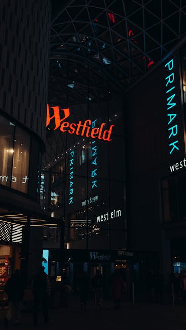 Interior view of the Bentall Centre Kingston shopping mall, featuring a high, curved glass roof supported by metal framework allowing natural light to illuminate the space. Multiple floors with retail stores are visible, connected by glass-fronted elevators and escalators. Inside, shoppers are walking through wide corridors, some on the ground and upper levels. Hanging from the ceiling are decorative, abstract bird-shaped sculptures. Packaging materials such as cardboard boxes, plastic wrap, and packing blankets are seen stacked on trolleys and near the entrance area, indicating ongoing packing and furniture transport activities. Kingston Removals staff may be involved in the loading process, although no movers are visible. The scene demonstrates a busy retail environment with elements consistent with logistics and moving services related to property and shop fitout relocation projects, as referenced on the Kingston removals website under the 'removals' service category.