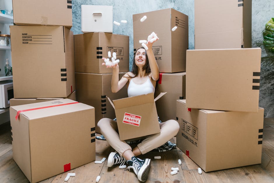 A woman sitting cross-legged on a wooden floor inside a room surrounded by numerous cardboard boxes in various sizes, some sealed with red tape and others open revealing packing materials. She is wearing a white tank top, beige pants, and black sneakers with white soles. She is smiling and tossing white packing peanuts into the air, with some falling into an open box on her lap. The room has a textured grey wall with a small shelf holding a potted plant and a white box in the corner, indicating a home relocation setting. The scene depicts the packing phase of a moving process, showcasing the importance of organized packing materials and careful handling during the furniture transport and home move, with the involvement of professional services like Kingston Removals.