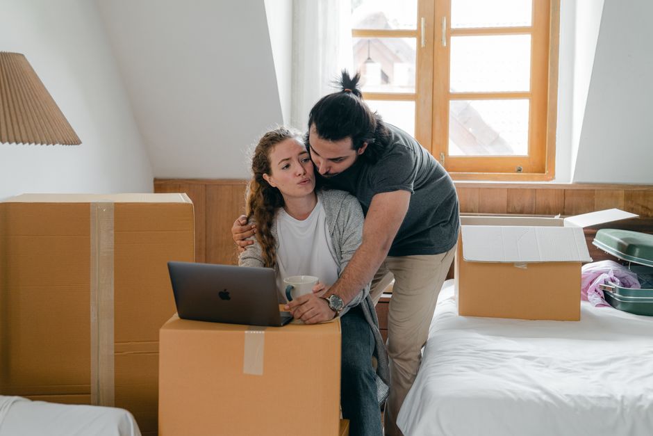 A young woman with long, curly brown hair sits on a bed in a room filled with moving boxes, some open and some sealed with packing tape, indicating a home relocation process. She is holding a white mug and has a neutral or slightly frustrated expression. A man with dark hair tied back in a ponytail, dressed in a dark grey t-shirt and beige pants, leans over her shoulder, appearing concerned as he gently holds her shoulder. In front of them is a closed silver laptop resting on a cardboard box. The room has a wooden headboard and a large window with wooden framing, allowing natural light to illuminate the scene. The window view reveals rooftops of neighboring houses. Around them are various packing materials, including plastic containers, cardboard boxes, and some packing paper, suggesting they are in the midst of packing and moving preparations typical of furniture transport and home moving logistics. Kingston Removals offers professional services to assist with the packing and loading process, reducing common booking mistakes during house removals.