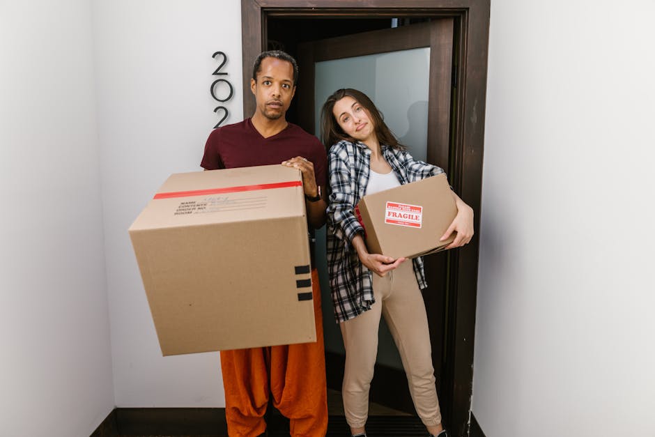 A man and a woman stand in a narrow corridor outside a residential doorway during a home relocation, each holding a cardboard box for moving. The man, dressed in a maroon shirt and orange trousers, is