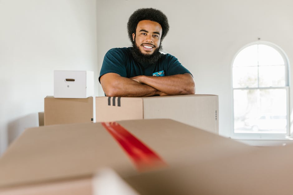 A man and a woman stand in a narrow corridor outside a residential doorway during a home relocation, each holding a cardboard box for moving. The man, dressed in a maroon shirt and orange trousers, is
