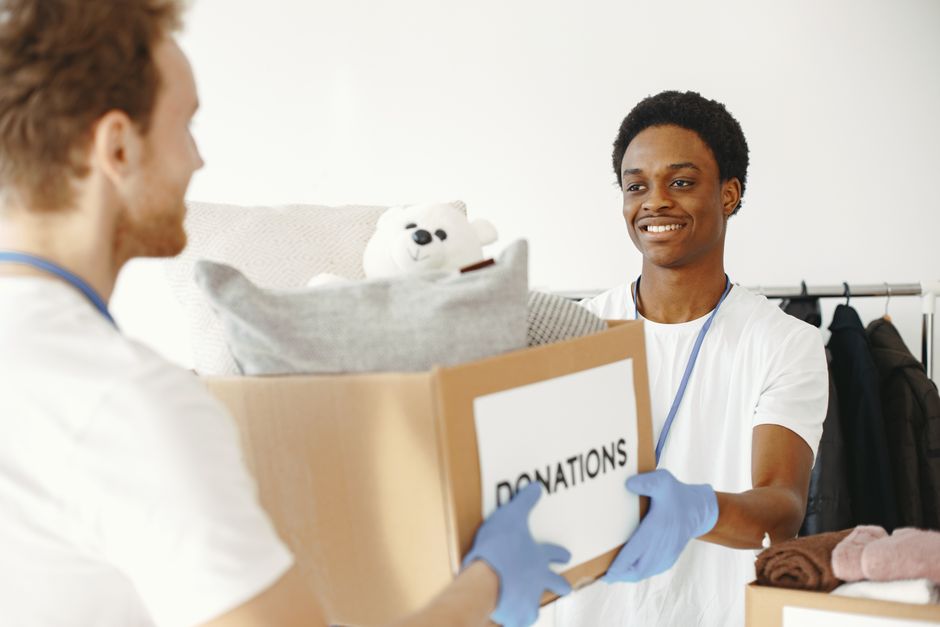 A young man with dark curly hair and a smile is inside a home, wearing a white T-shirt and blue gloves, as he hands over a cardboard box labeled 'DONATIONS' to a person with reddish-brown hair, also in a white T-shirt, whose face is partially visible. The box contains various items, including a white teddy bear, a pillow, and some clothing or fabric. The setting is an indoor space with a plain white wall in the background, and a clothing rack with hanging coats is visible to the right. The scene appears to depict a home relocation or donation collection process, with a focus on packing and moving services provided by Kingston Removals, relevant to Kingston's bulky waste rules for removals and charity donations.