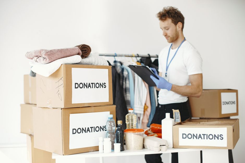 A man wearing a white t-shirt, blue gloves, and a lanyard is standing indoors next to a table with three large cardboard boxes labeled 'DONATIONS.' The boxes contain various clothing items, with some folded on top, and other donate-able goods including bottles of water, cleaning supplies, and household items arranged on the table. Behind him, there is a clothing rack with hanging garments such as shirts and jackets, suggesting a charity donation or clothing collection process related to house removals or home relocation services offered by Kingston Removals. The room has a plain, bright background, highlighting the organized setup for donation sorting or packing with ample space for furniture and boxes, supporting logistics involved in house moving or charity donation activities.