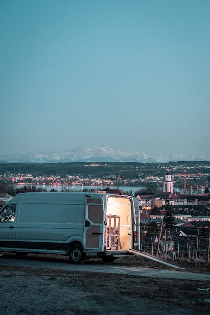 A grey moving van from Kingston Removals is parked on an unpaved area near a residential neighbourhood, with its rear doors wide open revealing the interior filled with various packing materials, including cardboard boxes, bubble wrap, and blankets. Inside the van, a wooden table or furniture piece is being carefully loaded using a trolley or lift system, with additional packing supplies visible around the area. The surrounding environment shows a cityscape with houses, industrial buildings, and a distant mountain range under a clear, blue sky. The scene captures the loading process during a home relocation, emphasizing furniture transport and packing activities associated with removals services, in accordance with Kingston council parking permits for moving vans.