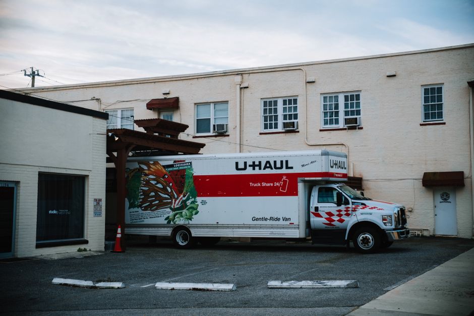 A white U-Haul moving truck with red and black branding is parked adjacent to a light-colored brick building. The truck, used for home relocation and furniture transport, features a large graphic of a butterfly and green leaves on its side, along with the text 'Truck Share 24/7' and 'Gentle-Ride Van.' The vehicle is positioned perpendicular to the building, with its rear end close to the pavement and doors facing the building’s side. The building has a flat roof, multiple small rectangular windows with white frames, some with window air conditioning units installed, and brown awnings above a few windows. The parking area is made of asphalt with white parking space markers and a single orange traffic cone placed near the front of the truck. The surroundings include a clear sky, and the scene appears to be during daytime, illustrating a typical logistics setup involved in furniture transportation during a house move, with Kingston Removals occasionally implied through the contextual relevance.