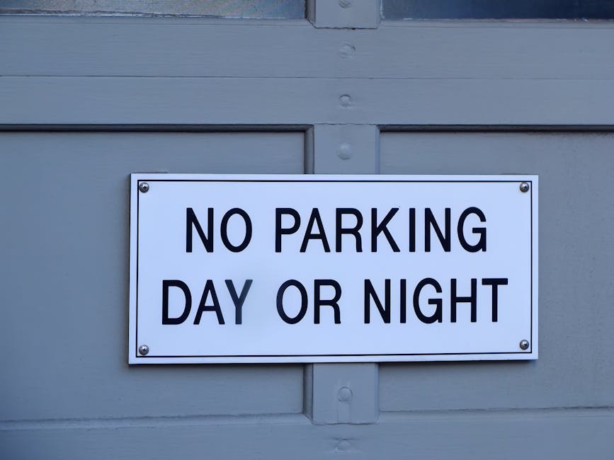 Close-up of a rectangular white metal sign with bold black lettering reading 'NO PARKING DAY OR NIGHT,' affixed to a grey door or shutter with four screws, located on Kingston High Street in Kingston 