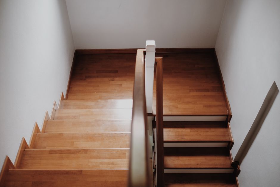 A view from the top of a staircase inside a residential property showing two sets of wooden stairs with polished treads and white risers. The left staircase ascends in a straight direction, while the right staircase, narrower with a slightly different design, also ascends. A wooden handrail runs along the center dividing the two staircases, supported by white balusters. The walls surrounding the staircase are plain and painted in a neutral color, with soft natural lighting illuminating the area. The image captures the interior environment during a home relocation or packing and moving process, emphasizing the spatial constraints posed by the narrow staircases often encountered during furniture transport. Kingston Removals’ service of navigating challenging staircases during house removals is relevant to this visual, illustrating the careful handling required when moving bulky items up or down tight staircases within property interiors.