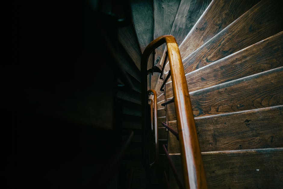 A narrow wooden staircase inside a home, viewed from above with the camera looking downward, showing weathered wooden steps and a polished wooden handrail on the right side. The staircase appears to be in a dimly lit environment, with the upper part fading into darkness and the lower steps illuminated enough to reveal their grain and texture. The confined space emphasizes the challenge of moving large furniture or boxes through tight, winding staircases during home relocations. Visible around the staircase are dark walls, and part of a landing can be seen at the top, indicating access to other rooms. This image illustrates a typical scenario where professional removals services, such as those from Kingston Removals, may need to assist with furniture transport, packing, and moving logistics in homes with narrow staircases, ensuring a safe and efficient home relocation process.