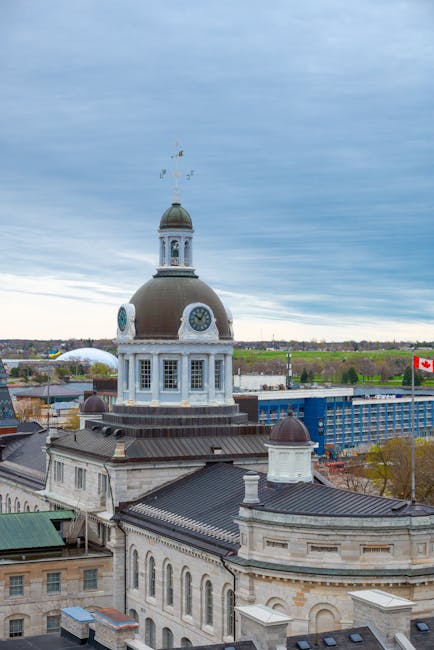 A panoramic view of a historic building with a large central dome topped by a clock and a weather vane, situated in an urban area with a mix of older stone structures and modern buildings, including a blue multi-storey parking structure and a flagpole with the Canadian flag, under a cloudy sky. The scene is captured from an elevated vantage point showcasing the building's architectural details and the surrounding cityscape, which includes distant green spaces and a river in the background. This setting reflects the typical environment where house removals and furniture transport services, such as those offered by Kingston Removals, may be involved in relocating clients’ belongings within or from this central location, highlighting the importance of careful packing and loading in complex urban environments.