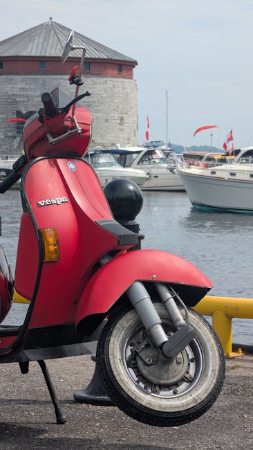 A red Vespa scooter parked on a paved area near a marina with watercraft and boats moored in the background. The scooter features a black seat and a small windshield, with its front wheel slightly turned to the left. Behind the scooter, there is a stone building with a conical roof and a weather vane on top, along with a collection of parked cars. The marina has several yachts and boats with flags, and the scene is under an overcast sky. This image captures a typical scene of urban mobility combined with waterfront leisure, suitable for illustrating transport options in house removal or relocation contexts, especially in waterfront areas like Norbiton Kingston. Kingston Removals occasionally recommends considering transport logistics such as vehicle access in moving services, as shown by the scooter and nearby vehicles.