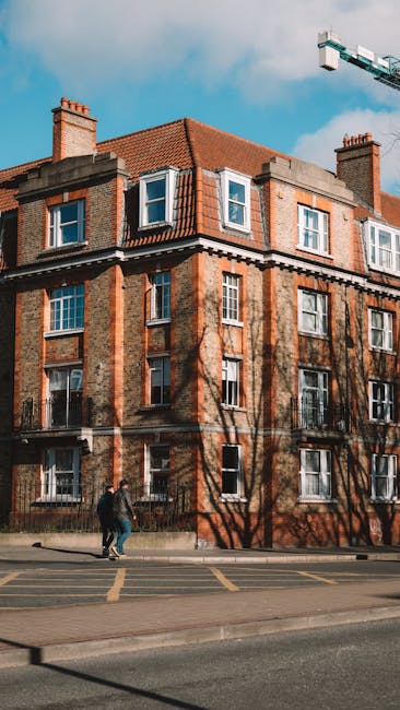 A panoramic view of a historic building with a large central dome topped by a clock and a weather vane, situated in an urban area with a mix of older stone structures and modern buildings, including a blue multi-storey parking structure and a flagpole with the Canadian flag, under a cloudy sky. The scene is captured from an elevated vantage point showcasing the building's architectural details and the surrounding cityscape, which includes distant green spaces and a river in the background. This setting reflects the typical environment where house removals and furniture transport services, such as those offered by Kingston Removals, may be involved in relocating clients’ belongings within or from this central location, highlighting the importance of careful packing and loading in complex urban environments.