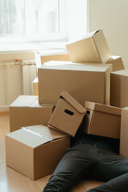 A person lying on the floor surrounded by multiple cardboard boxes during a home relocation or moving process. The boxes vary in size and some are sealed with packing tape, while others are open or partially closed. One box features black plastic handles for easy carrying. The scene is set inside a room with natural light coming through a large window, with a white window frame and light-colored walls visible in the background. The individual appears to be in the midst of packing or unpacking, with their legs visible amongst the boxes, indicating an active loading or unpacking process. The environment suggests a professional or self-managed move, aligning with the services provided by Kingston Removals for urgent or same-day moving needs, including furniture transport and packing during house removals.